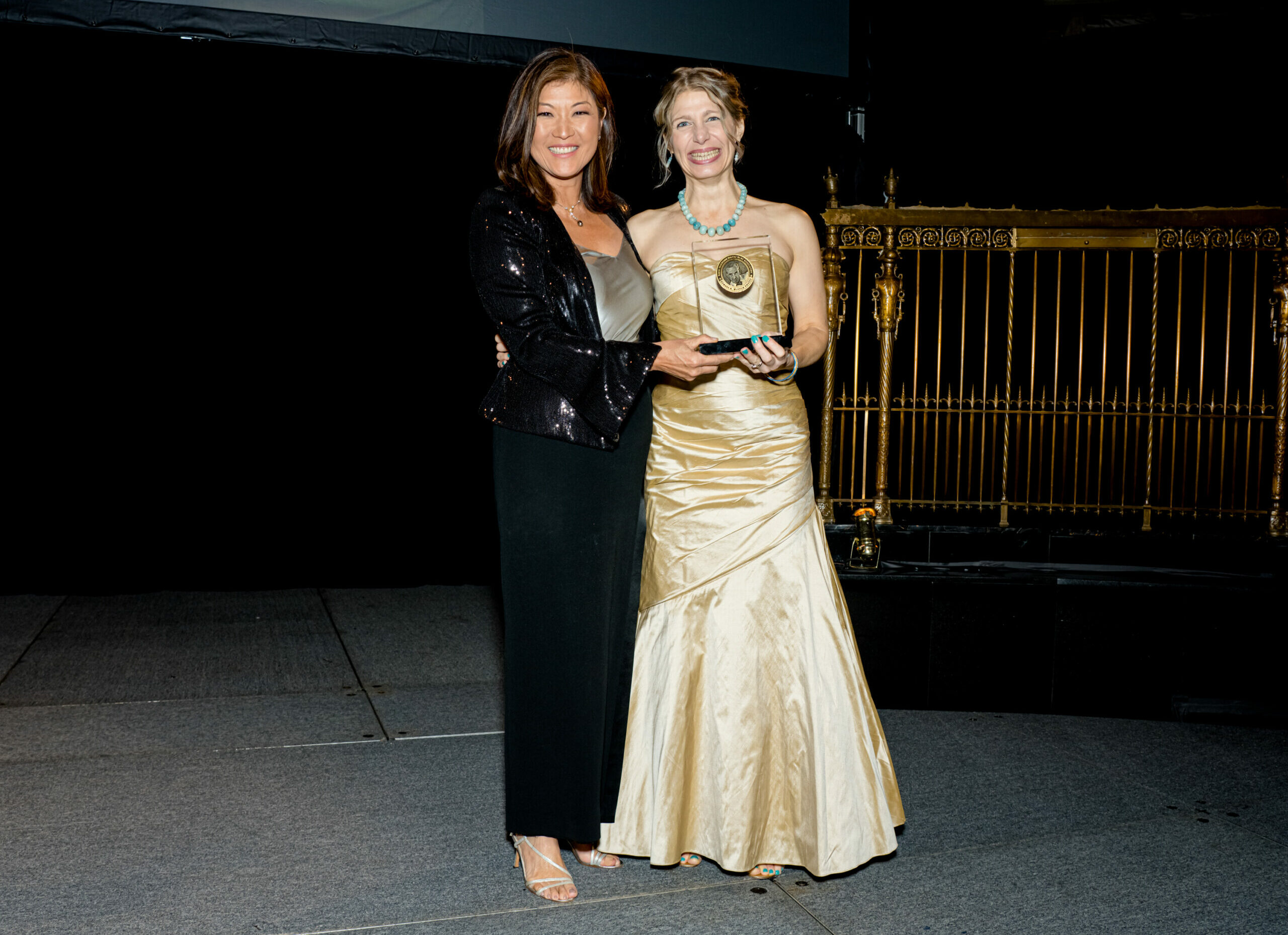 Two smiling women in formal dresses holding an award